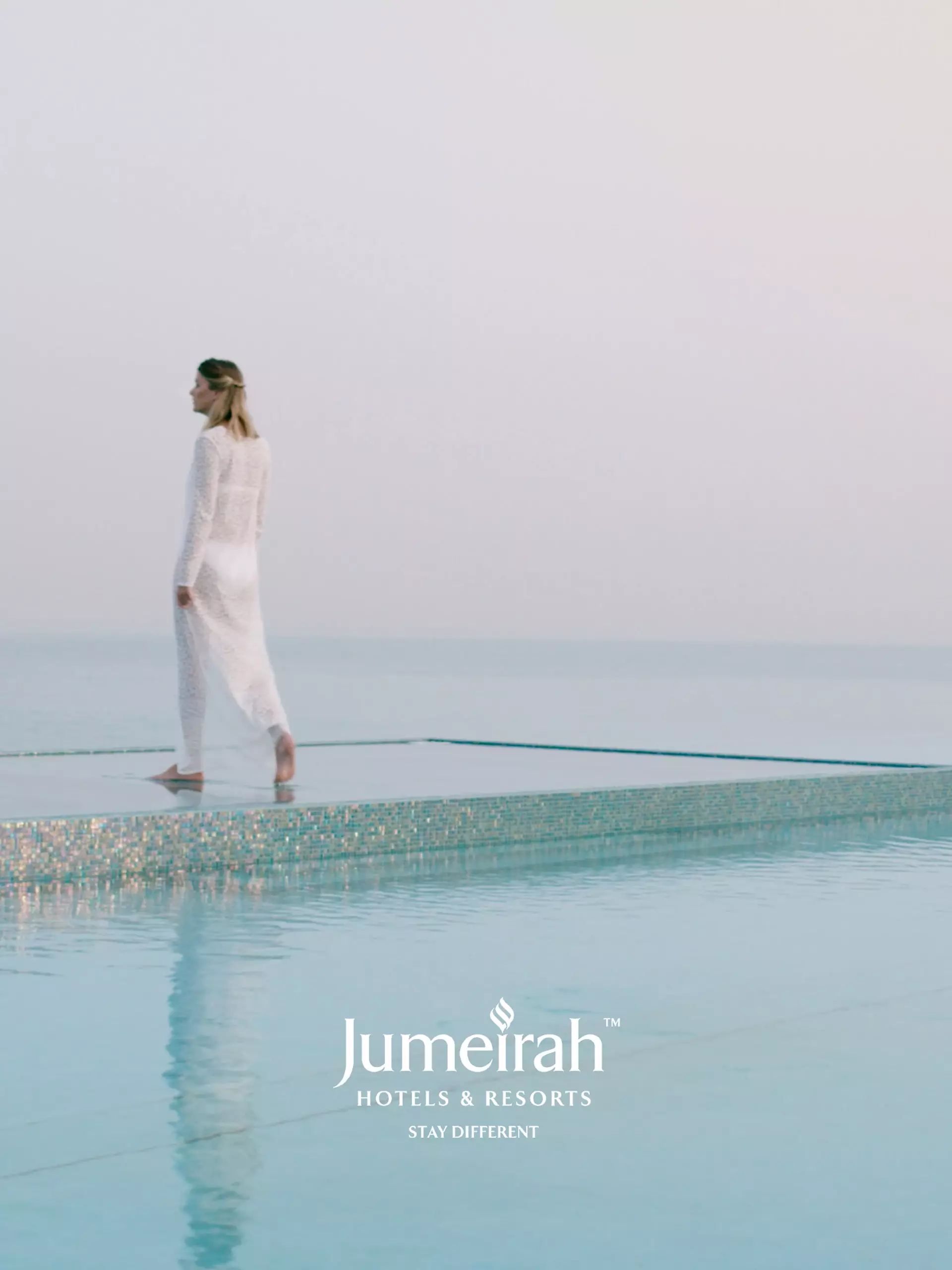 Frau in weißem Kleid geht am Rand eines Infinity-Pools mit ruhigem Meer im Hintergrund, Jumeirah Hotels & Resorts Logo