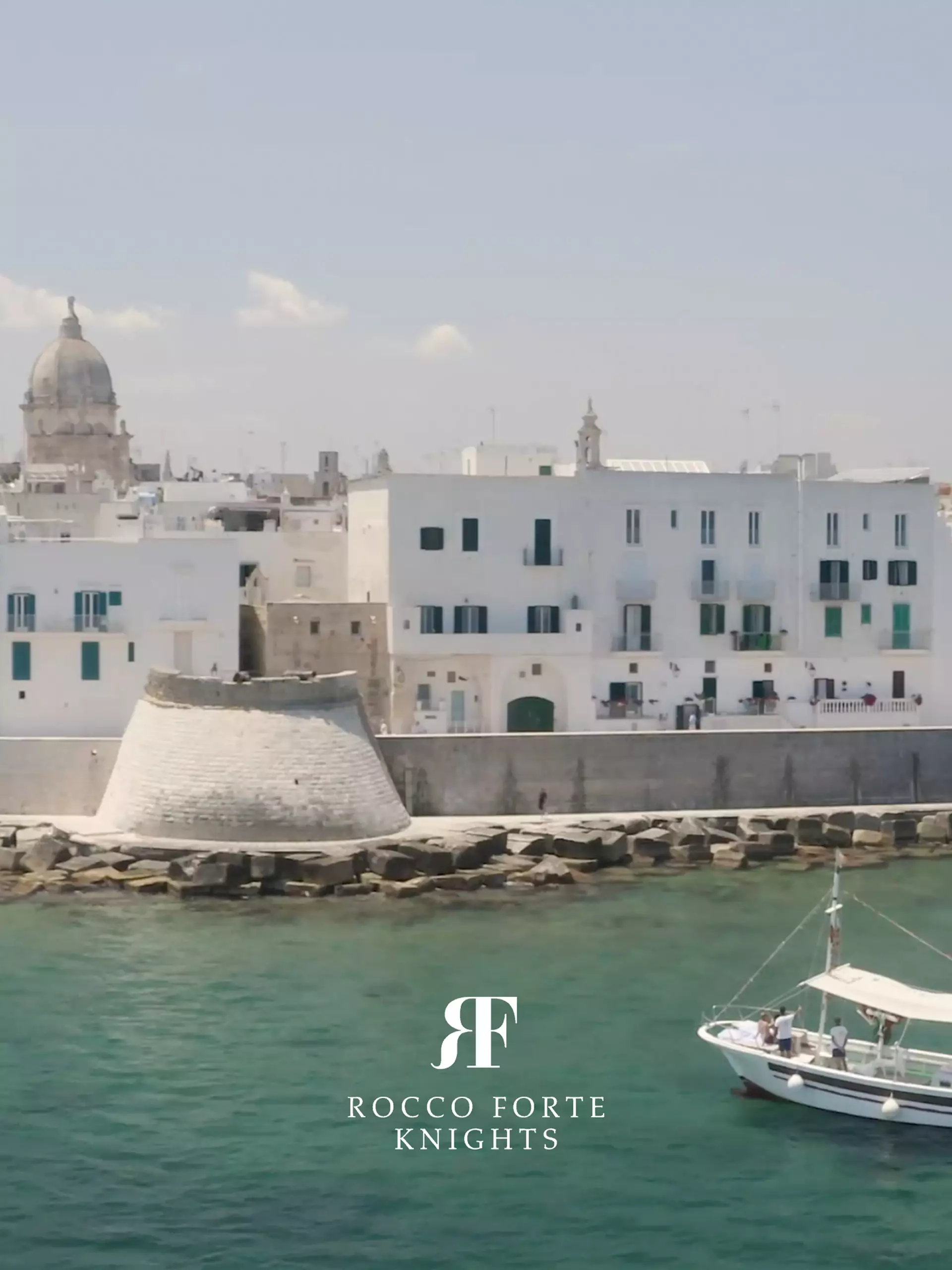Küstenstadt mit weißer Festungsmauer und Boot auf klarem Wasser, Logo Rocco Forte Knights eingeblendet