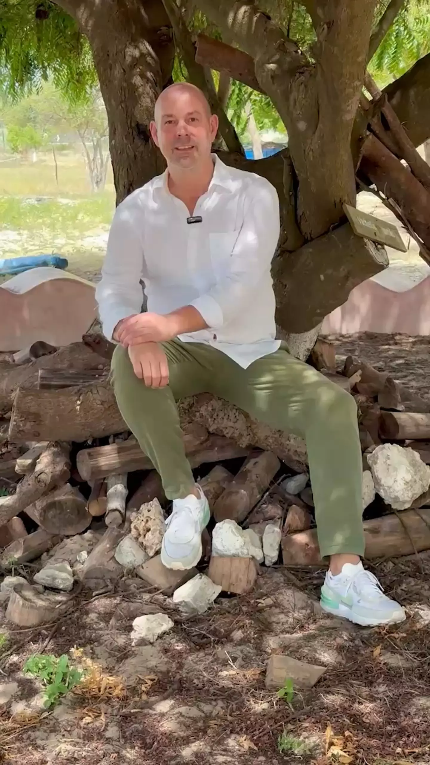 Mann in weißem Hemd und grüner Hose sitzt auf Holzstapel unter Baum im Freien, symbolisiert Engagement vor Ort in Kenia
